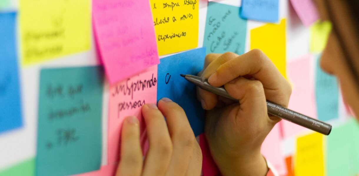 Manos escribiendo un mensaje en una nota adhesiva rosa, rodeada de más notas de colores pegadas en la pared
