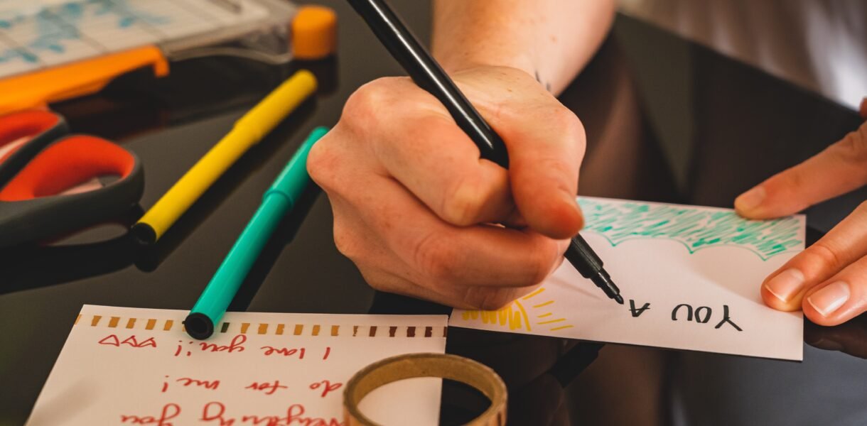 Manos escribiendo una tarjeta a mano con marcador, rodeada de tijeras, marcadores de colores y cinta adhesiva