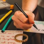 Manos escribiendo una tarjeta a mano con marcador, rodeada de tijeras, marcadores de colores y cinta adhesiva