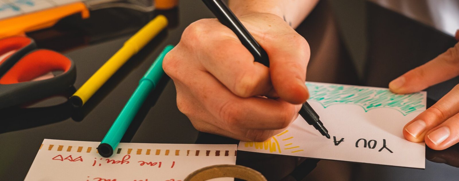 Manos escribiendo una tarjeta a mano con marcador, rodeada de tijeras, marcadores de colores y cinta adhesiva